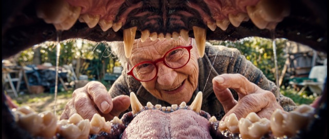 An elderly woman in red glasses peering into the open jaws of a giant creature